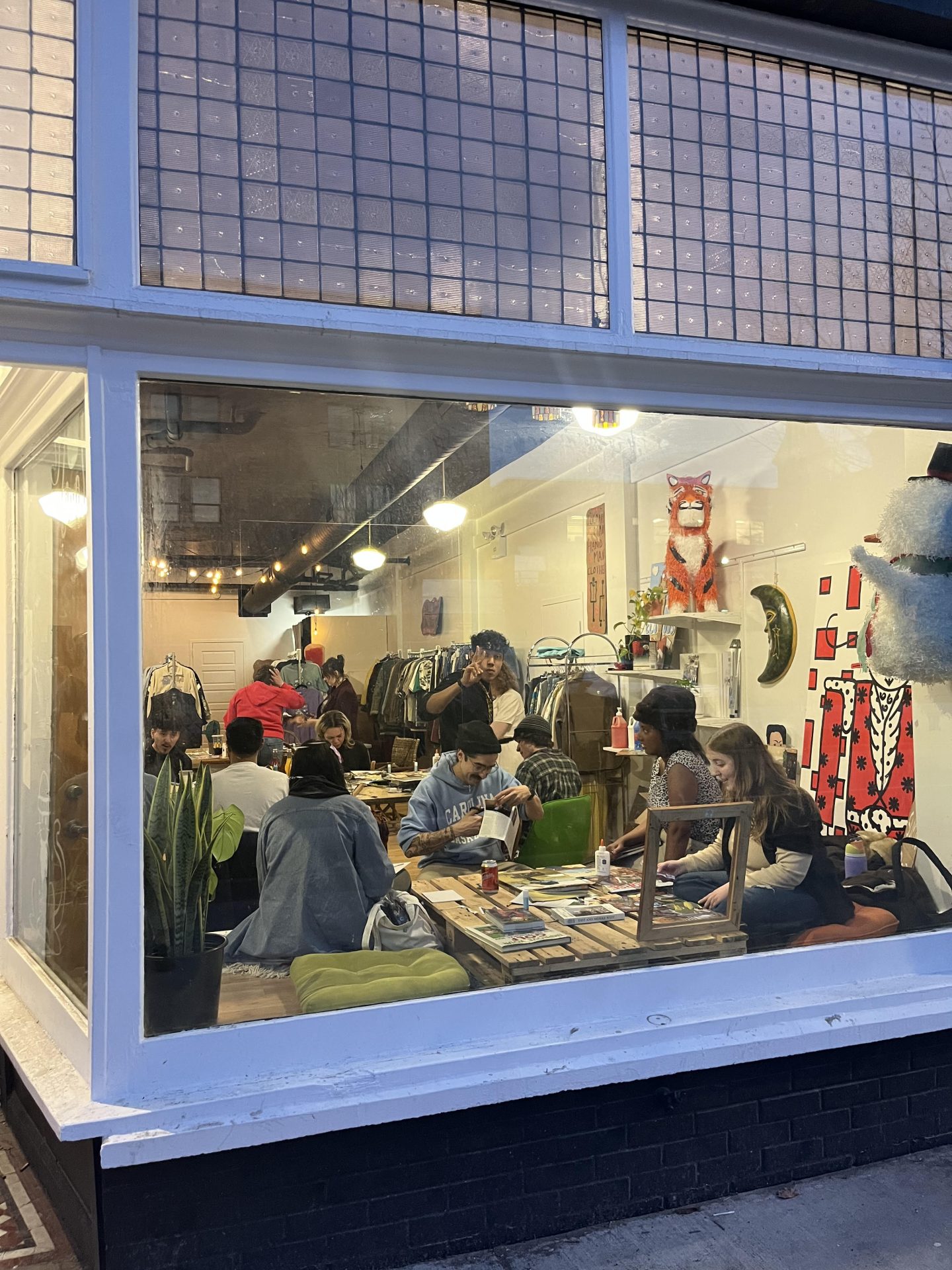 A view of NIDO's front window at evening. Inside, tables of young people work on canvases as part of a group art project.